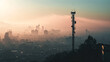 Early morning view of a cell tower silhouetted against a hazy city skyline as the sun rises, casting a soft glow.

