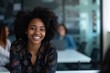 © Régis Cardoso - young woman sitting at a table in an office with other people in the background. She is smiling and looks happy.