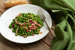 A white plate is filled with vibrant green peas and crispy pieces of bacon, accompanied by a silver fork on the table next to it.