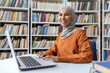 A cheerful young woman wearing a hijab sits at a library table, using her laptop among rows of books. Her smile indicates confidence and contentment with her study environment.