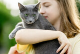 Fototapeta Koty - Cute teenage girl holding her pet cat on sunny summer day. Gorgeous grey Russian Blue breed cat outdoors.