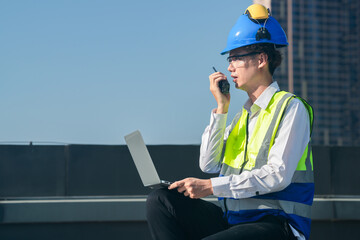 Focused engineer in hard hat and safety vest using walkie-talkie and laptop on construction site with urban backdrop.