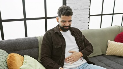 Wall Mural - Young latin man suffering from painful stomachache at home, hand on his belly depicts unwell feeling of illness. expression of stomach pain, nausea suggestive of disease.