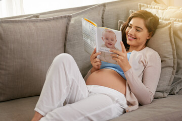 Poster - Happy and pregnant woman, reading baby book at home to prepare for motherhood on sofa. Mother, smile and learning to parent with novel or story for education and research to relax before childbirth