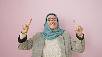 Sticker - Cheerful middle age hispanic woman in hijab pointing upwards, smiling in surprise and joy over the pink isolated background, her finger highlighting the space above in amazement.