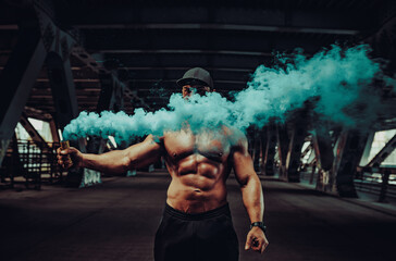 Wall Mural - Young strong man bodybuilder standing with dense blue smoke on urban industrial background