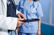 Close-up shot of a caucasian doctor in a lab coat in a clinic office using a smart tablet. Pair of male hands holding a digital device for medical research while a nurse waits in the background.
