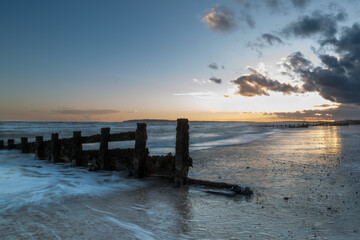 Wall Mural - Long exposure landscape photograph at Camber sands beach on a warm evening, Image shows the beautiful sunset and glowing sky with a damaged wooden groyne subject and a receding tide