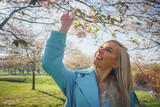 Fototapeta Miasta - Summer fashion portrait of pretty blonde woman posing on amazing blooming Sakura tree background. Happy travel woman smile near a sakura cherry blossoms tree