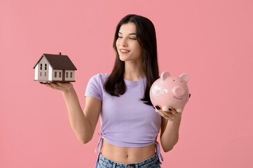 Wall Mural - Beautiful young happy woman with piggy bank and house model on pink background. Mortgage concept