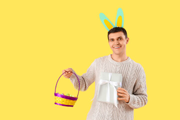 Canvas Print - Happy young man in bunny ears with gift box and wicker basket on yellow background. Easter celebration