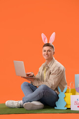 Sticker - Young man in bunny ears with laptop shopping on grass against orange background. Easter Sale