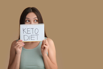 Poster - Beautiful young thoughtful Asian woman holding notebook with text KETO DIET on brown background