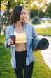 Adult beautiful smiling woman with cup of coffee and roll mat, standing in the park. Take a break.