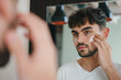 Handsome young man applying cream to his face while looking in the mirror, taking great care of his skin.