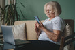 Senior woman using smartphone and laptop computer working online sitting on sofa indoors. Senior professional texting via cellphone. Browsing internet.