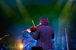 A violinist musician stands on stage with a violin in his hands during a concert. There is a bright light from the floodlights around.