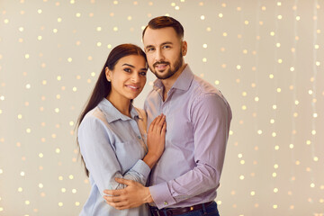 Portrait of a young happy smiling man hugging brunette girl standing indoors on lights bokeh background and looking cheerful at camera. Happy couple in love. Relationships and Valentines day concept.