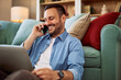 A happy adult man talking on a phone while leaning against a couch behind him.