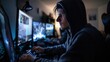 Young man in hoodie working late on computer in dark room with glowing screen at night time