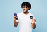 Fototapeta  - Young Indian man wear white t-shirt casual clothes using mobile cell phone hold credit bank card do online shopping order delivery book tour isolated on plain blue cyan background. Lifestyle concept.