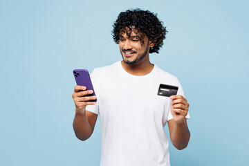 Young Indian man wear white t-shirt casual clothes using mobile cell phone hold credit bank card do online shopping order delivery book tour isolated on plain blue cyan background. Lifestyle concept.