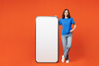 © ViDi Studio - Full body young fun woman wear blue t-shirt casual clothes stand near big huge blank screen mobile cell phone smartphone with workspace copy space mockup area isolated on plain red orange background.