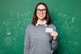 Fototapeta  - Young happy smart teacher woman wear grey casual shirt glasses hold credit bank card isolated on plain green wall white chalk blackboard background studio. Education in high school college concept.