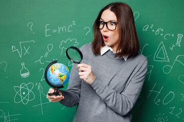 Wall Mural - Young shocked sad smart teacher woman wear grey casual shirt glasses hold globe use reading-glass isolated on green wall chalk blackboard background studio. Education in high school college concept.