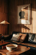A dark home office interior with decor on a wooden table in front of a black leather sofa with two pillows. A sconce, and a vertical poster with a wooden frame hanging on a wall with wooden panels.