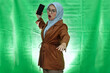 angry young Asian woman wearing hijab, glasses and blazer with displeased expression and wanna throw mobile phone over green background
