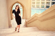 Beautiful sensual young woman with long dark hair in black dress walking on Dubai Downtown street.