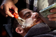 Working in Barbershop. Professional Barber applying foam on customer's cheeks for shaving.