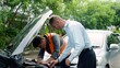 Middle-aged male in light blue shirt peers into car engine while mechanic in orange vest works, both focused on issue. Concept of taking care of car insurance after an accident.