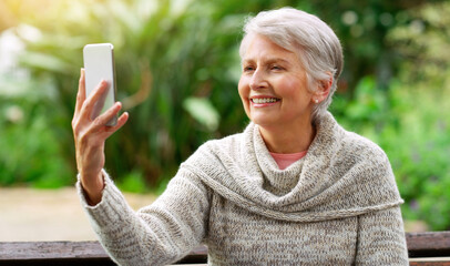 Poster - Senior woman, phone and selfie in park for social media post, travel and relax on garden bench. Female person, smile and camera app for picture in outdoor nature, photo and holiday in retirement