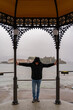 middle aged man wearing a black windbreaker posing under a gazebo by the lake on a rainy day - travel and tourism concept