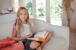 a little girl 6-7 years old is getting ready for school, the girl is dressed in a light-colored top and a plaid skirt.  The kid lsit on the sofa  with a backpack and a book