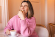 Happy woman eating cookie sitting in a restaurant. Positive woman bites cookie enjoys eating sweet food breaks diet closed eyes. Woman wear pink sweater rest in cafe. Girl bite cake and look happy.