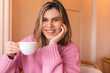 Portrait of gorgeous smiling lady hold and drinking cappuccino from cup while resting in restaurant. Girl laughing and look at camera. Happy woman wear pink sweater. Listen with interest.