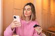 Calm young woman wear pink knit sweater clothes with credit card in hand using mobile phone paying for purchase online relaxing sitting at table in cafe. Serious blonde girl.