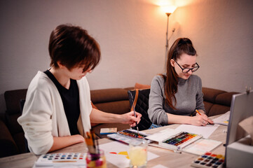 Two concentrated women involved in an artistic project, with one painting and the other measuring, surrounded by creative tools