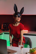 A beautiful girl in a bdsm-style rabbit mask and a bright red dress with leather straps is posing sweetly in the kitchen in neon light