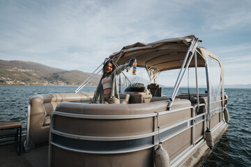 Wall Mural - A happy, youthful woman raises her drink in celebration while standing on a pontoon boat, surrounded by a scenic lake view.