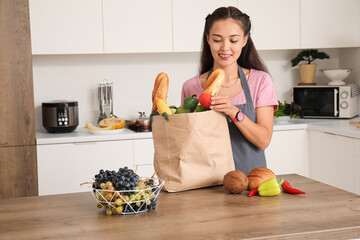 Poster - Beautiful young Asian woman with grapes and paper bag full of fresh products in kitchen