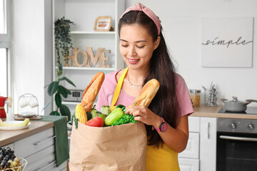 Poster - Beautiful young Asian woman with paper bag full of fresh products in kitchen