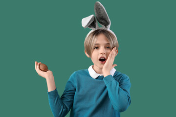 Poster - Shocked little boy in bunny ears with chocolate egg on green background. Easter celebration