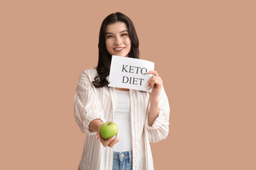 Wall Mural - Happy smiling young woman holding paper sheet with text KETO DIET and apple on beige background