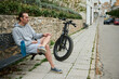 © Taras Grebinets - Young man cyclist sitting on a bench near his electric bike, relaxing after ride in mountain city