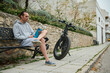 © Taras Grebinets - Young adult sportsman, athletic man cyclist relaxing when riding electric bike in the city, holding a bottle of water while sitting on the bench. Active people. Urban lifestyle. Eco-friendly transport