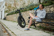© Taras Grebinets - Full length portrait cyclist man drinking water, sitting on city bench. Electric motorbike, e-bike, electric bicycle bike on the foreground. Bike sharing city service. Sustainability.